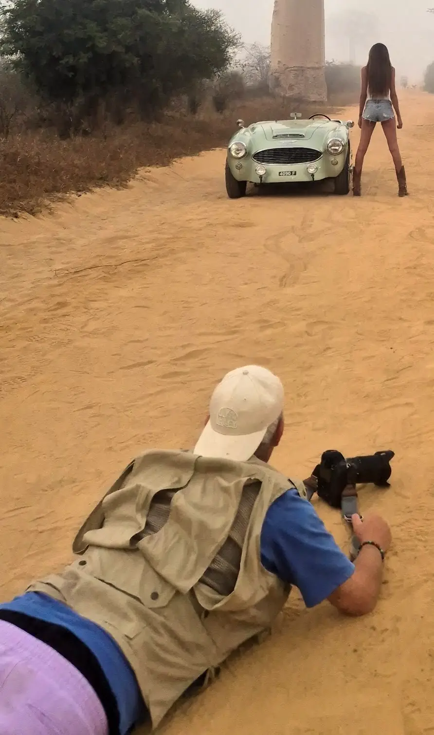 Photographe allongé sur le sable photographiant un mannequin et un roadster ancien sur l’Allée des Baobabs, Madagascar