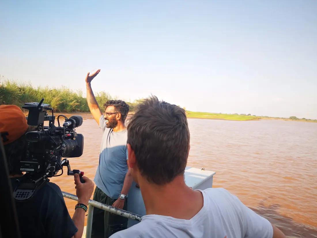 Presenter waving from a boat on a wide red river while the crew films